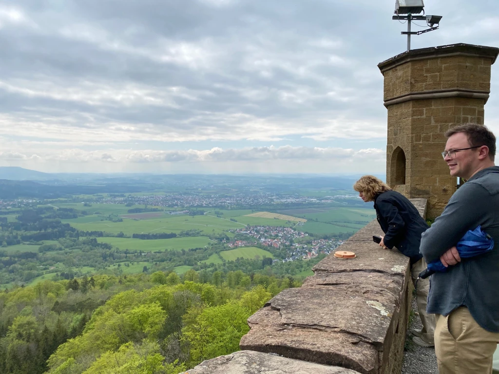 Vince & Jack Croyle stand on a stone wall, savoring the view of green fields and scattered houses beneath a cloudy sky. One leans gently while the other, with a backpack slung over his shoulder, stands by—moments like these remind them that family matters most.