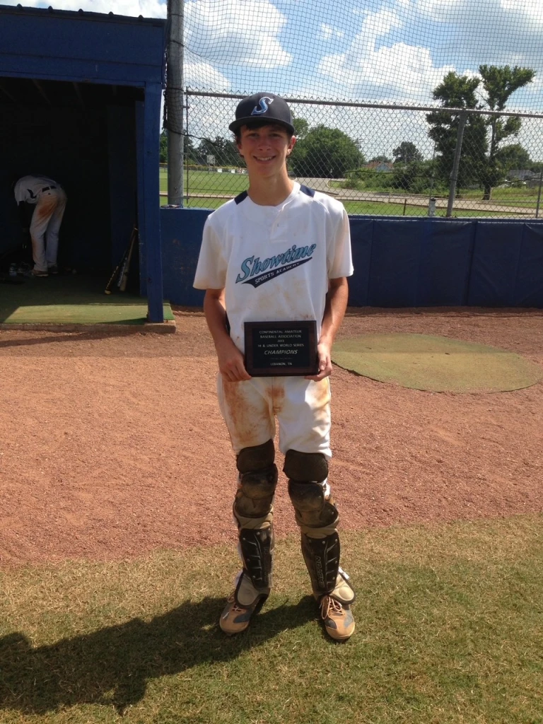 A young baseball player, possibly Jack Croyle, wearing a white and blue uniform and cap, stands on the baseball field holding a plaque. The player is in catcher's gear with dirt marks on the uniform. A dugout and another player are visible in the background.