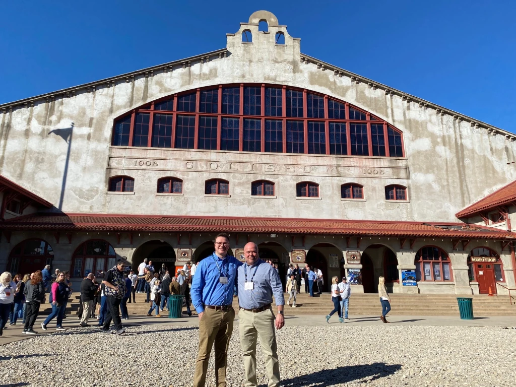 Two men, possibly Jack and Vince Croyle, stand in front of an old brick building with large arched windows and a sign reading "1908." People walk around in the background under a clear, blue sky. It feels like a scene from "Family Matters," where history and community intertwine seamlessly.