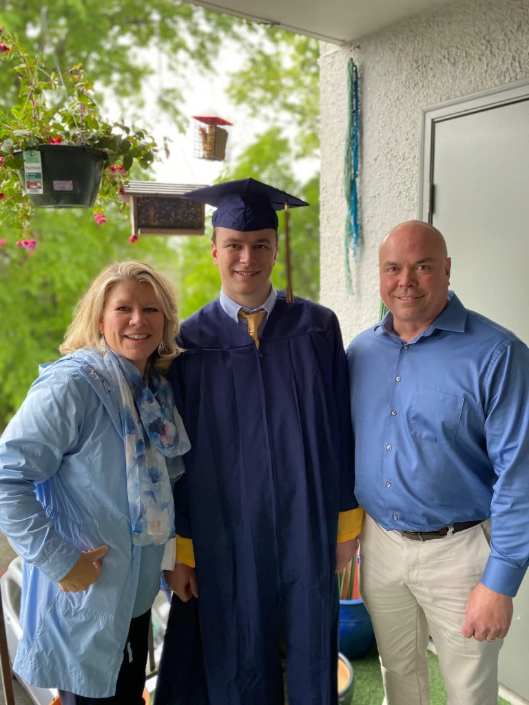 A graduate in a blue cap and gown stands between two adults, a woman on the left and Vince Croyle on the right. They are smiling on a balcony with greenery in the background. The woman wears a light blue scarf, and Vince sports a blue shirt. It's truly a Family Matters moment.