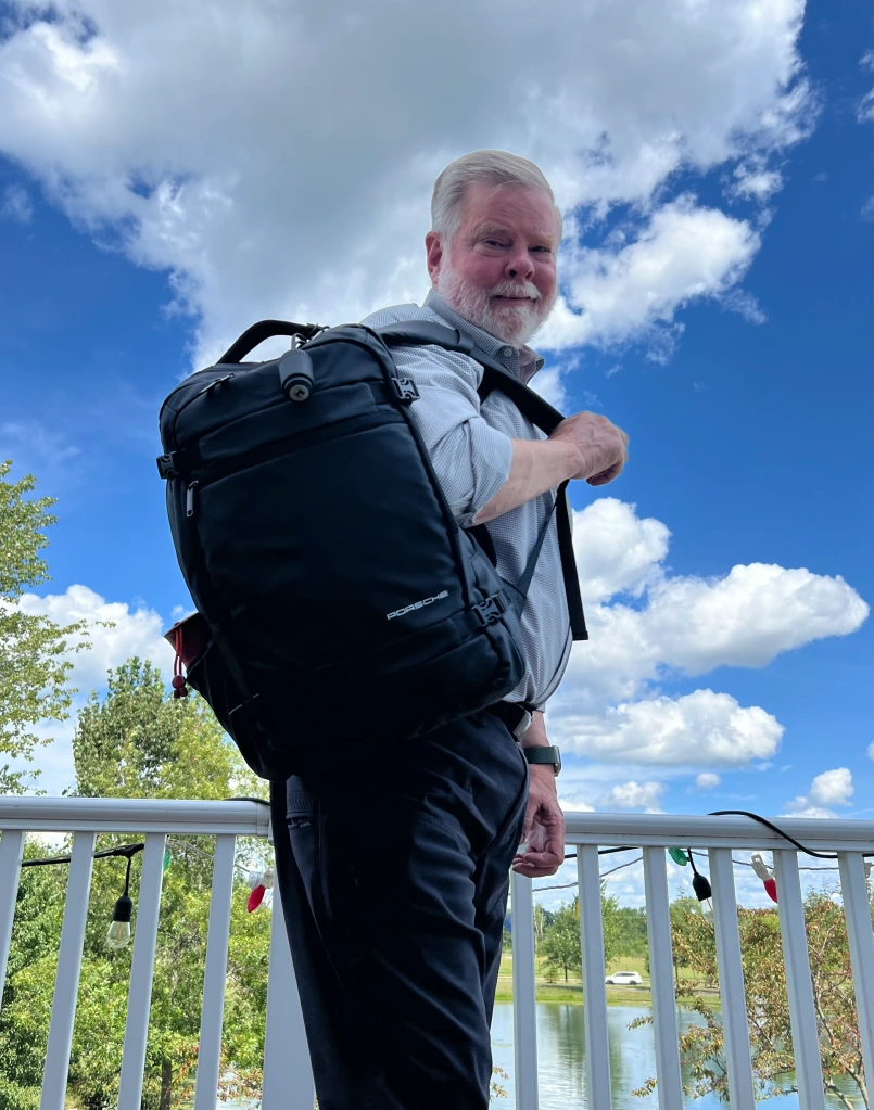A person with a white beard stands outdoors on a balcony on August 20th, 2024, carrying a black backpack. The sky is partly cloudy, and trees and a body of water are visible in the background, making it feel like a serene break from the typical work day.