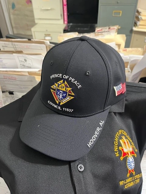 A black cap and shirt are placed on a chair, ideal promotional products for Work Day. The cap flaunts a "Prince of Peace" emblem with "Council 11537," while the shirt boasts a Knights of Columbus design. Both pieces, embroidered with "Hoover, AL," sit against a backdrop of file folders and cabinets.
