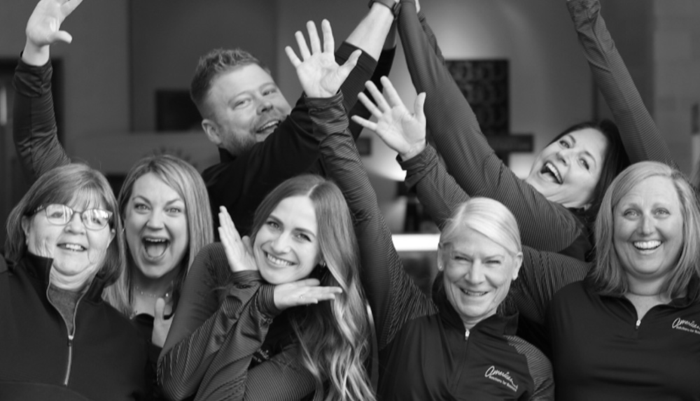 A group of seven smiling people pose enthusiastically with arms up, some making playful gestures, embodying a vibrant culture. They are wearing casual jackets and shirts. The black and white photo beautifully highlights their joyful expressions.
