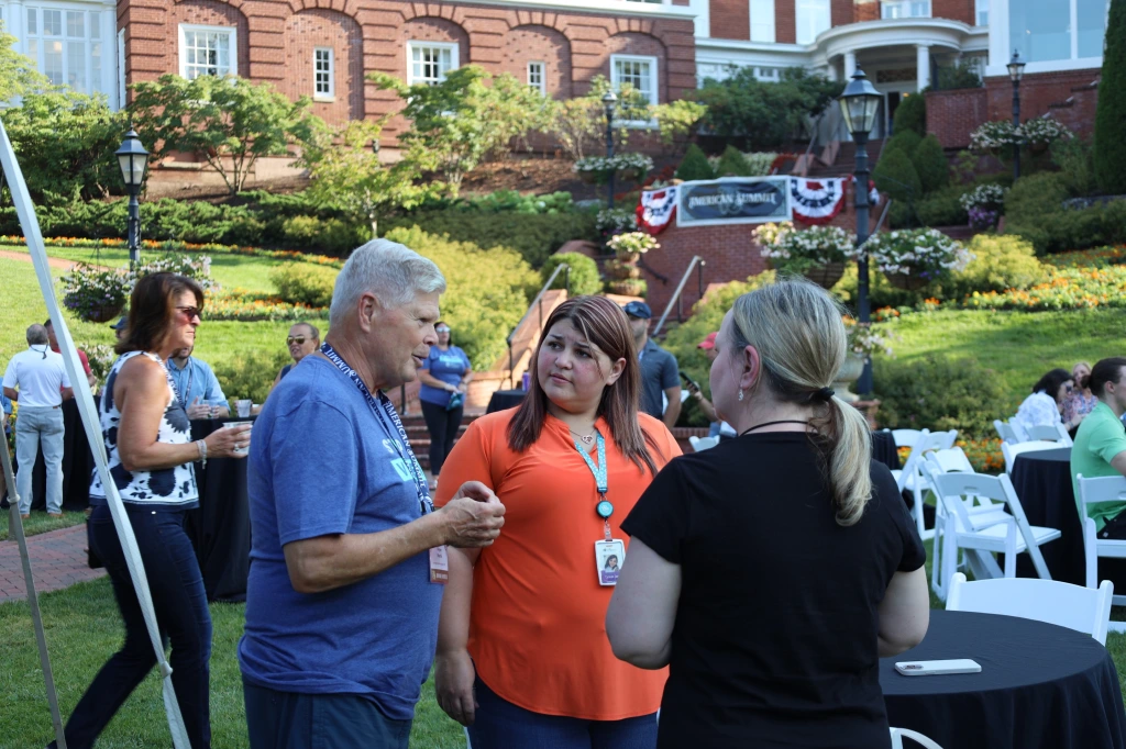 A group of people socialize outdoors at a daytime event hosted by Safehome Systems. Tables are set up on a grassy area with buildings and greenery in the background. Attendees engage in conversation, savoring the gathering under clear weather and discussing innovative security solutions.