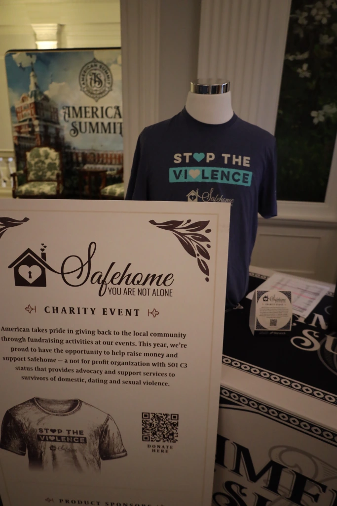 A mannequin donning a "Stop the Violence" t-shirt stands behind a charity event sign for Safehome Systems, an organization supporting domestic and sexual violence survivors. In the background, "American Summit" is partially visible, highlighting the event's connection to ASB fundraising efforts.