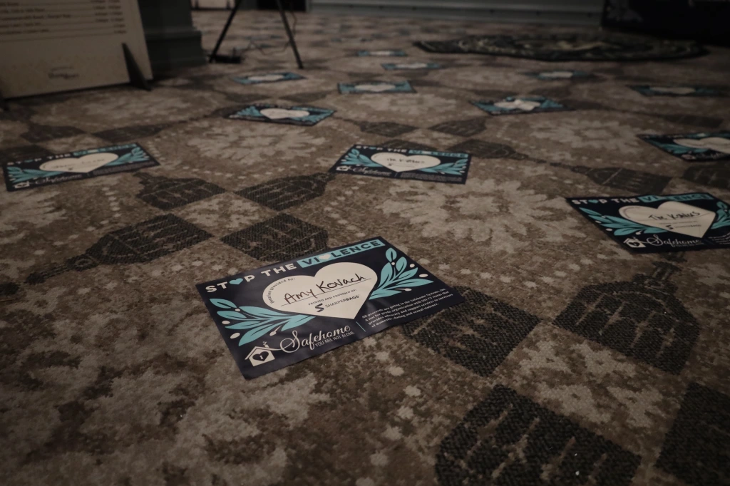 The floor is adorned with a patterned carpet, scattered with name tags displaying a blue heart and the words "Stop the Violence." One tag reads "Amy Koukalos," hinting at a fundraising effort by ASB in support of Safehome Systems.