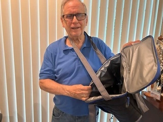 An elderly man wearing glasses and a blue shirt stands indoors, holding an open gray bag with a shoulder strap, perfect for storing promotional products. Vertical blinds in the background let in soft light, adding to this scene's cozy ambiance, captured on August 20th, 2024.