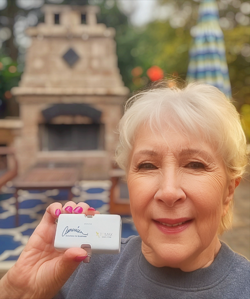 An elderly woman with short white hair smiles while holding a small white box with a signature and logo, showcasing delightful promotional products. She stands outdoors in front of a brick fireplace, a rug, and an umbrella on this sunny August 20th, 2024.