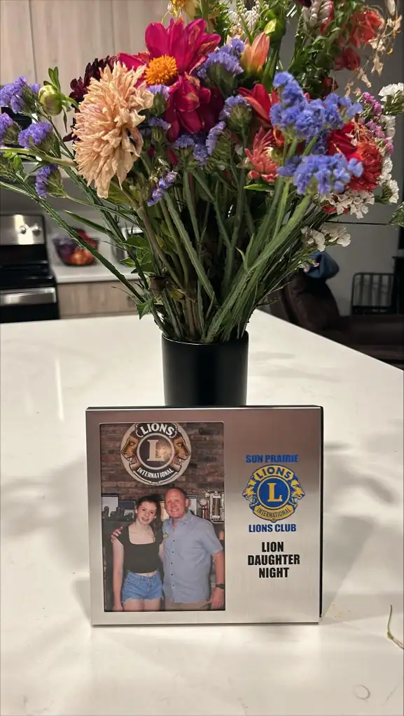 A colorful bouquet of mixed flowers in a black vase graces a white countertop. In front, a framed photo captures two people at Sun Prairie Lions Club's "Lion Daughter Night." The frame, adorned with logos and text, evokes the spirit of the club's memorable events like August 20th, 2024.