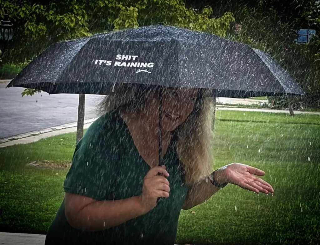 A woman stands under a black umbrella with "SHIT IT'S RAINING" printed in white. Smiling, she holds out her hand as rain pours down, set against a backdrop of a green lawn and trees. This quirky promotional product perfectly captures the spirit of unexpected August 20th showers.