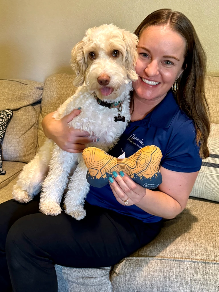 A woman sits on a couch, smiling and holding a white, fluffy dog in her arms. She’s also playfully showcasing a bone-shaped toy with a trendy wood grain pattern—a thoughtful detail of our latest promotional products collection launching August 20th, 2024. Beige cushions and part of a decorative pillow enhance the cozy ambiance.