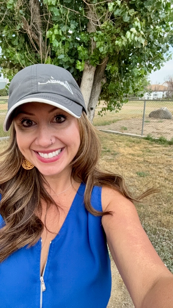 A woman in a blue sleeveless top and gray cap is smiling at the camera. On this sunny August 2024 day, she's outdoors in front of a tree and fence, with grassy fields stretching behind her—a perfect backdrop for capturing promotional product photos.