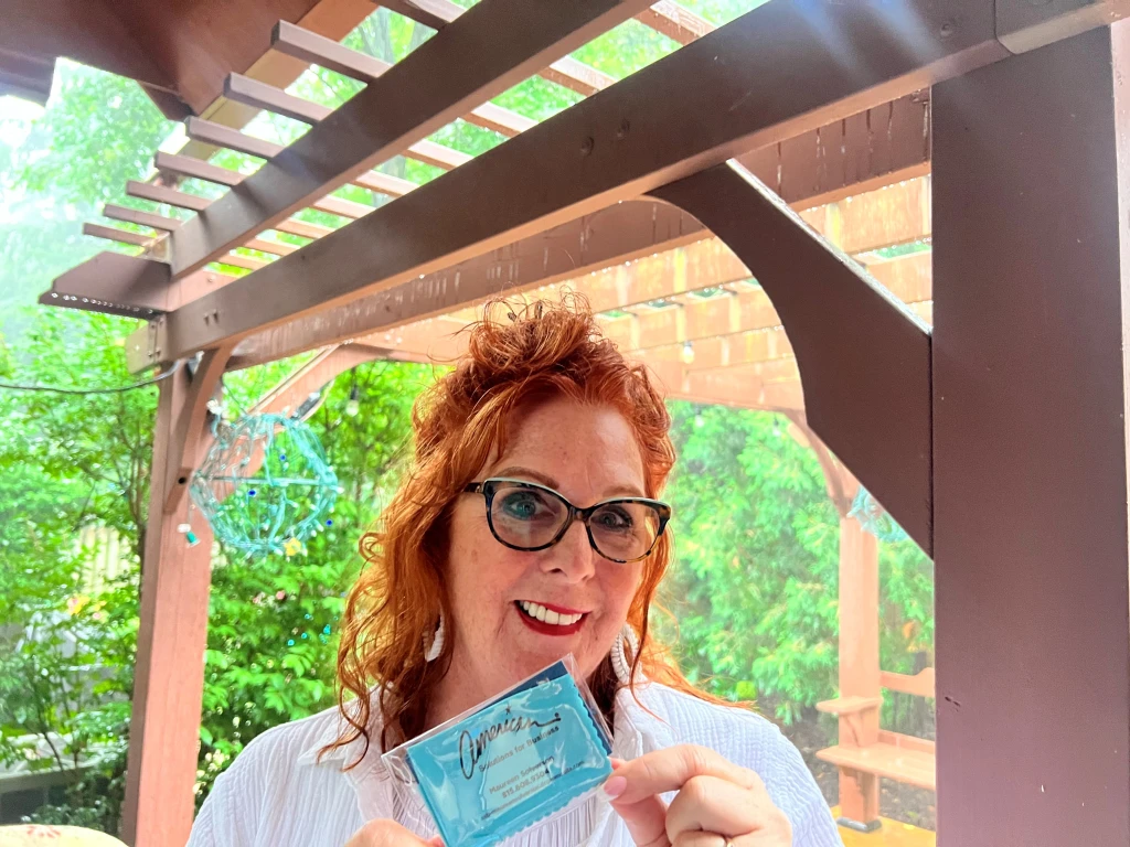 A smiling person with red hair and glasses proudly holds up a blue card, like one of those innovative promotional products, under a wooden pergola. Lush greenery paints the background like an August dreamscape.