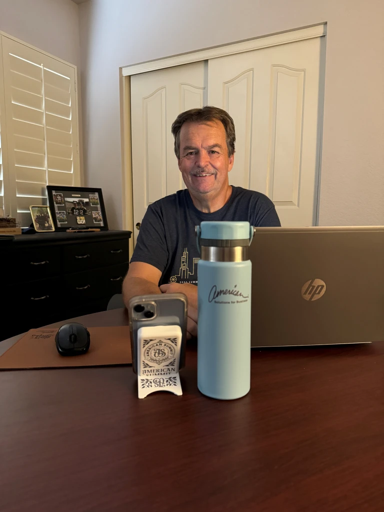 A man sits smiling at a desk with a laptop, mouse, smartphone on a stand, and a blue promotional water bottle. A framed picture and closed doors are in the background, capturing the essence of Promotional Products Work Day as we look forward to August 20th, 2024.