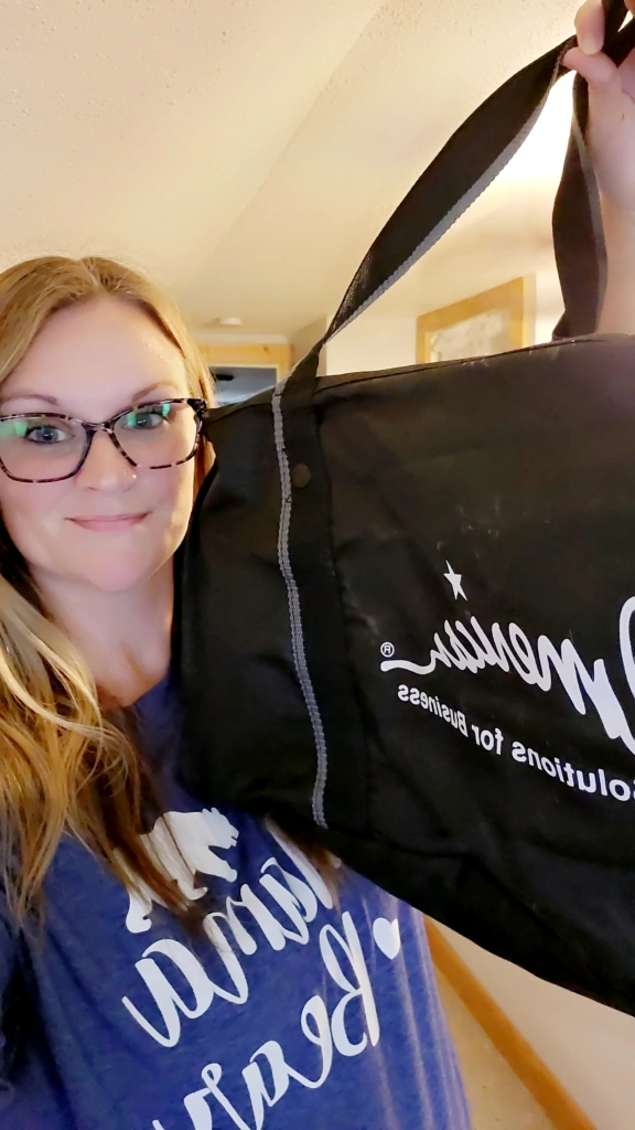 A person with glasses smiles, clutching a black bag with "Solutions for Business" in white text—an ideal promotional product for any work day. They're wearing a blue shirt adorned with cursive writing. The backdrop is a softly lit room, capturing the essence of August 20th, 2024.
