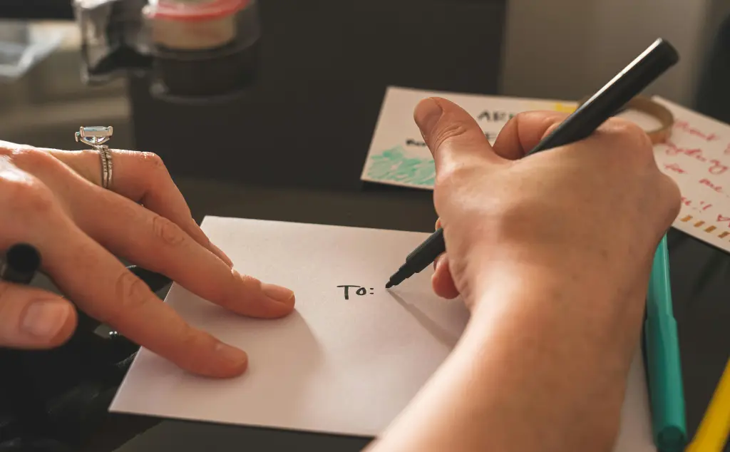 A person is writing "To:" on a white envelope with a black marker, engaging in the timeless practice of direct mail. The envelope rests on a dark surface, surrounded by colorful pens and other stationery items. Another hand steadies the envelope in this simple yet meaningful act amidst our digital world.