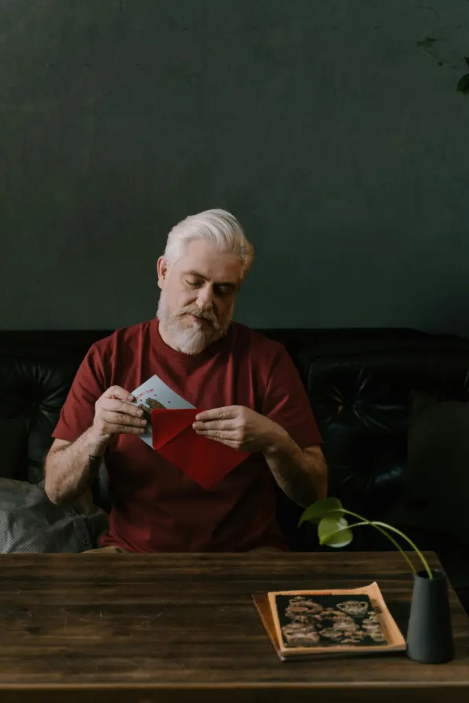 An elderly man with white hair and a beard sits on a dark couch, concentrating intently as he opens a red envelope. A small green plant and a magazine rest on the wooden table before him, casting soft shadows in the dimly lit room.