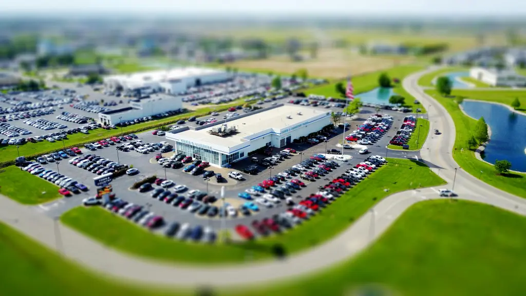 From an aerial view, the expansive auto dealership resembles a strategic draft plan, with countless vehicles meticulously parked in rows around the central building. Roads and green spaces surround the lot, while distant buildings dot the horizon under a clear sky.