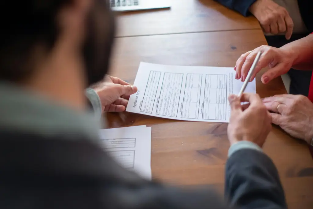 A person reviews a document with pen in hand at a wooden table, while another points at the paper, engaging in a discussion about business continuity. Their faces are obscured. Other papers and part of a laptop hint at surrounding dealership strategies.