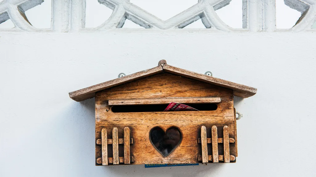 A wooden mailbox with a heart-shaped cutout is mounted on a white wall. With its roof-like top and fence-like details, the mailbox stands as a charming nod to direct mail amidst our digital world. A piece of mail is partially visible inside, waiting to be discovered.