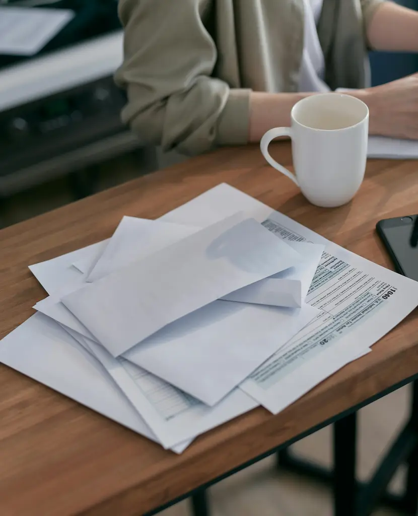 A person is seated at a wooden table with a stack of envelopes and papers, suggesting they might be organizing an auto draft setup. A white mug rests nearby, while the edge of a smartphone peeks through in this casual home or office setting.