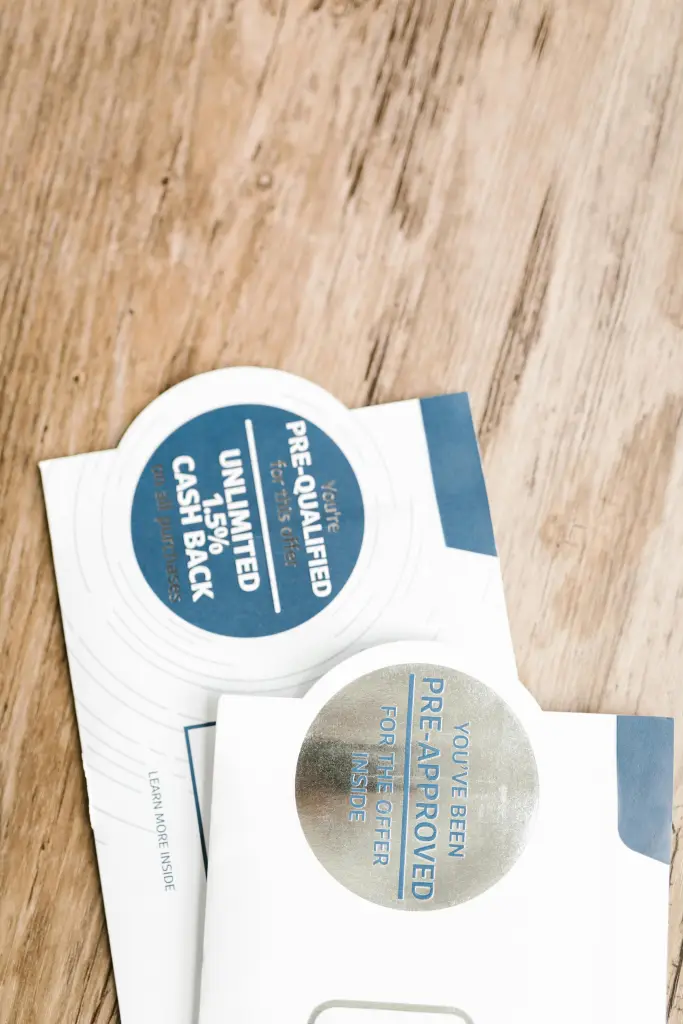 Two envelopes lay overlapping on a wooden surface, marked by circular stickers. One states "You've been pre-qualified" with cash back details, the other "You've been pre-approved." Both mirror the blue and white branding typical of financial institutions, blending seamlessly into the direct mail landscape.
