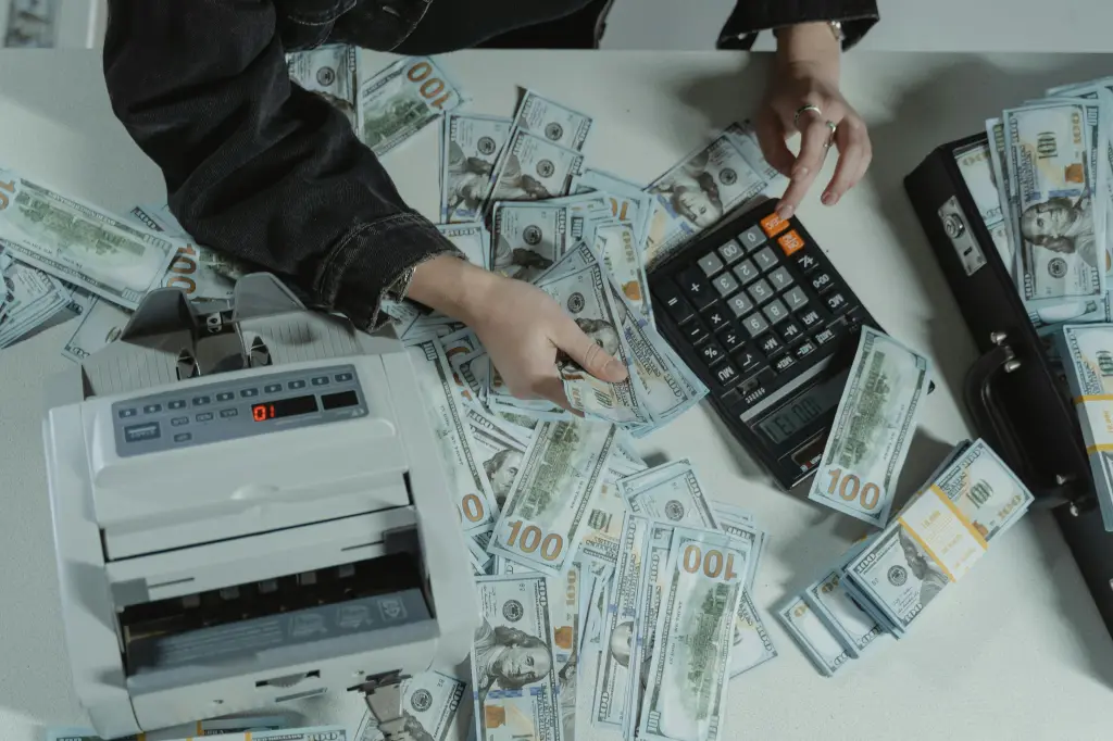 A person uses a calculator amid scattered US hundred-dollar bills on a table, with a money counting machine nearby. The hands are poised over the money, suggesting sorting or counting—a wake-up call for ensuring business continuity in this financial landscape.
