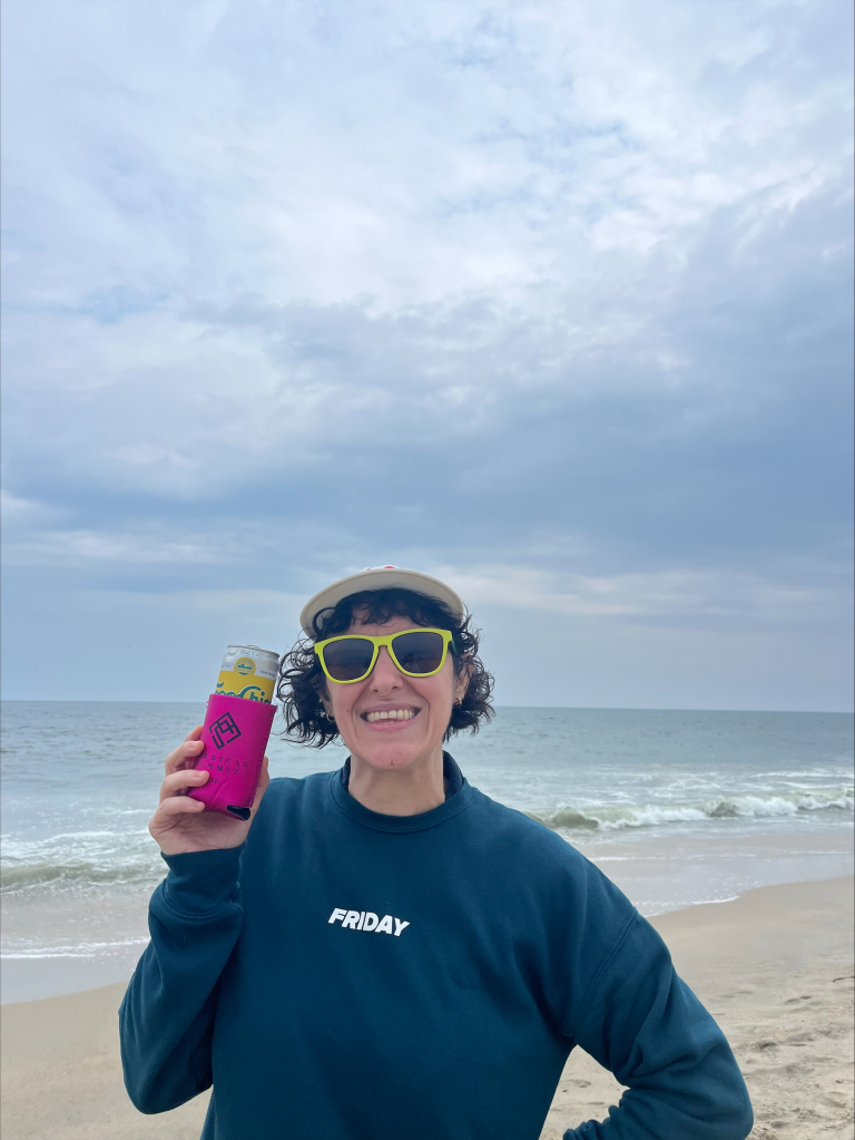 A person with curly hair and yellow sunglasses holds a promotional drink in a pink koozie, standing on a beach. They're smiling, wearing a cap and a blue sweatshirt that says "FRIDAY." The sky is cloudy, and waves crash in the background—a perfect escape from the typical work day.