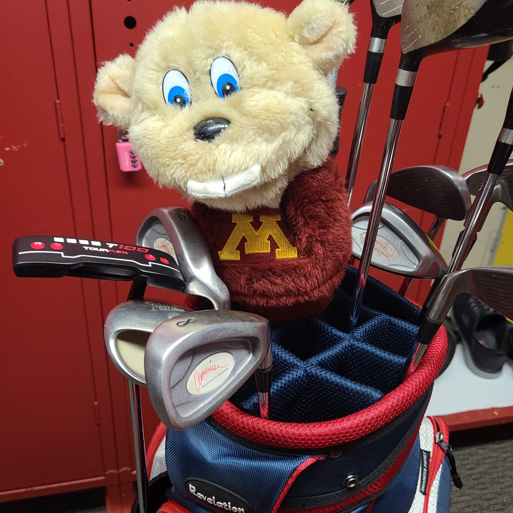 A golf bag filled with clubs features a plush toy headcover, styled as a smiling gopher in a red sweater with a yellow "M," perfectly blending fun into any work day. Ideal for promotional products, this cheerful accessory stands out against the backdrop of red lockers.