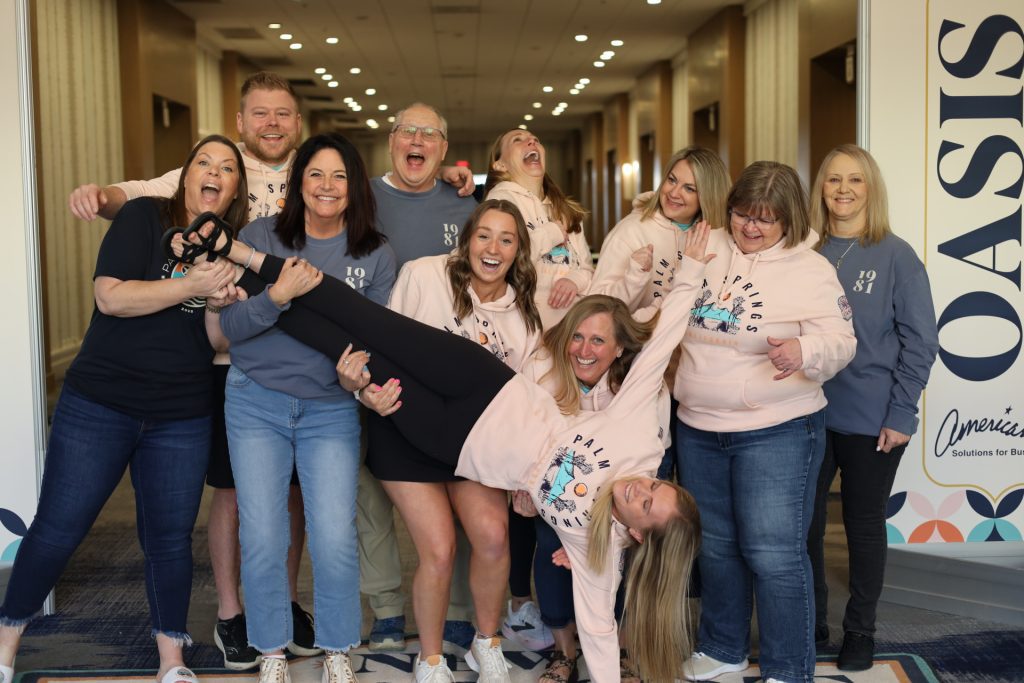 At the lively National Sales Conference in Palm Springs, a joyful group of nine poses on a blue-carpeted hallway. Laughter fills the air as two people lift one woman horizontally. Clad in casual, colorful attire, this playful crew radiates energy and camaraderie.