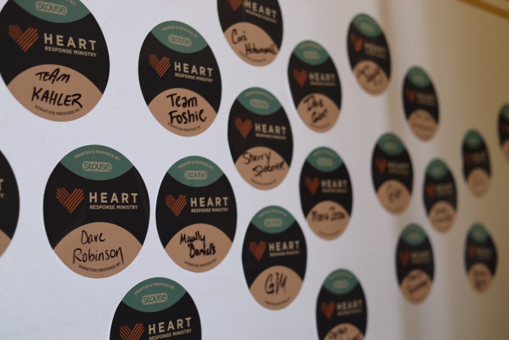 A whiteboard in Palm Springs boasts numerous round stickers from the Heart Response Ministry. Each sticker, linked to the National Sales Conference, is labeled with various handwritten names and teams, primarily featuring colors in black, white, and red.