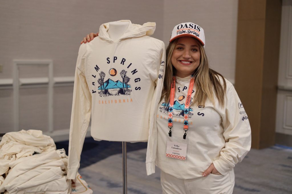 A woman in a Palm Springs-themed outfit poses next to a mannequin at the National Sales Conference. Both sport hoodies showcasing CA's iconic desert scenery. Her cap proudly declares "Oasis Palm Springs.