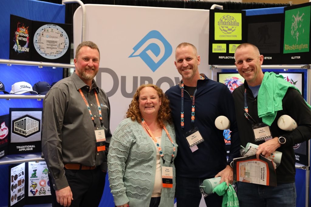 Four people are standing and smiling in front of a Dupont display booth at the National Sales Conference. They appear to be enjoying the event in Palm Springs, holding promotional items while the backdrop showcases various graphics and logos.