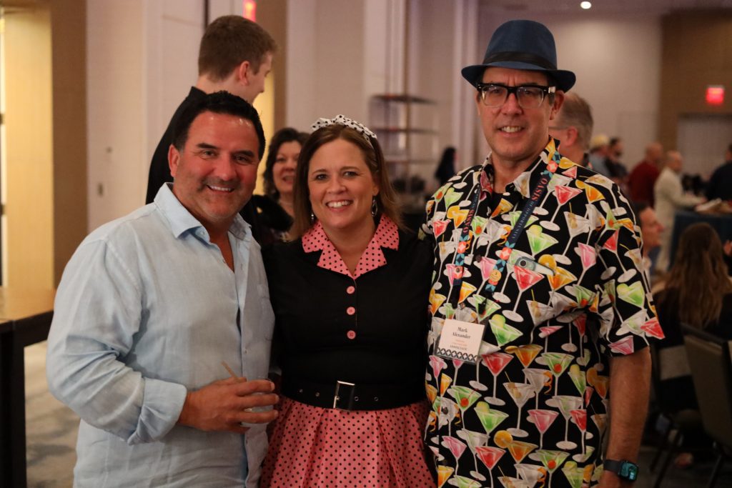 Three people are posing together indoors at a National Sales Conference in Palm Springs. The woman in the center dons a tiara and pink polka dot dress. ASB Hosts' energetic vibe shines through as the man on the right wears a hat and cocktail-themed shirt, while the one on the left sports a light blue shirt.
