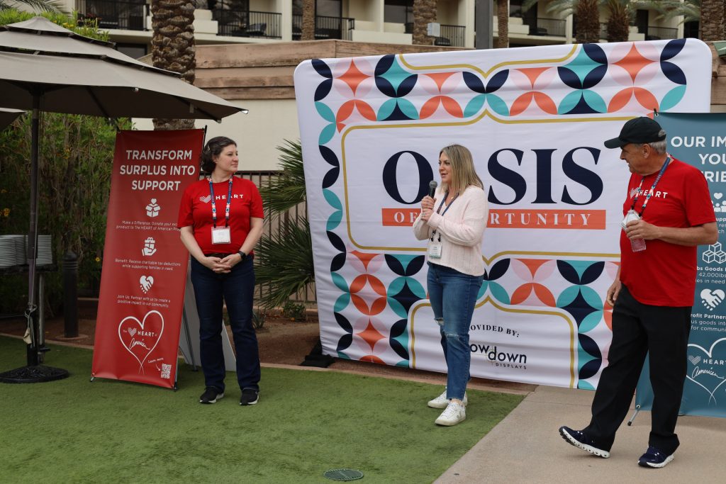 A woman speaks into a microphone beside a banner with "Oasis of Opportunity" in large letters. At this charity event, two people in red shirts listen intently—one stands near an umbrella-shaded table, as a sign reads "Transform Surplus Into Support.