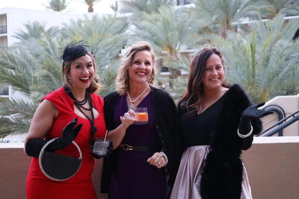 Three women dressed in elegant attire stand on a Palm Springs balcony during the National Sales Conference. One wears a red dress with black gloves, the second a purple dress with a necklace, and the third in black with a tiara. They're smiling and holding drinks as palm trees sway in the background.
