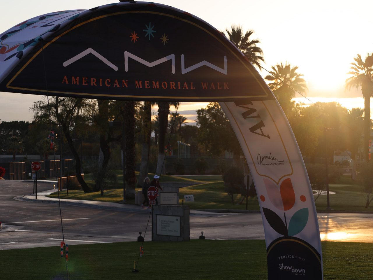 A large archway over a field reads "American Memorial Walk" with logos and designs. As the sun sets, casting a warm glow over palm trees and the grassy area, it feels reminiscent of a serene venue for charity events or even an inspiring National Sales Conference.