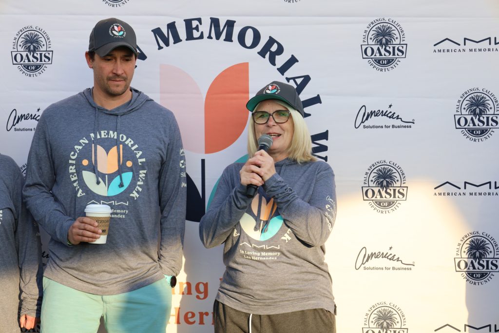 A woman with glasses and a cap speaks into a microphone at a charity event, while a man beside her holds a cup. Both are in matching American Memorial Walk shirts. The backdrop, adorned with logos, hints at the esteemed ASB's involvement in the gathering.