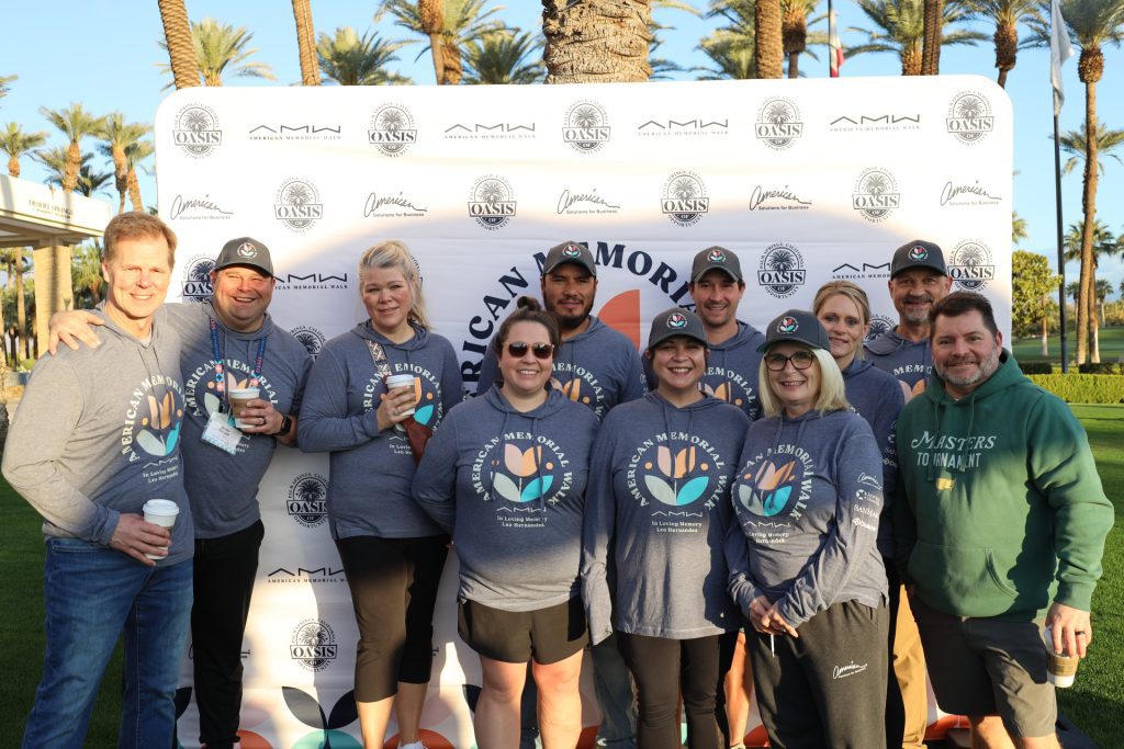 A group of people wearing matching blue event shirts and holding drinks pose in front of a banner outdoors. Palm trees sway in the background, hinting at the relaxed vibe of the National Sales Conference. They are smiling, clearly enjoying this casual gathering.