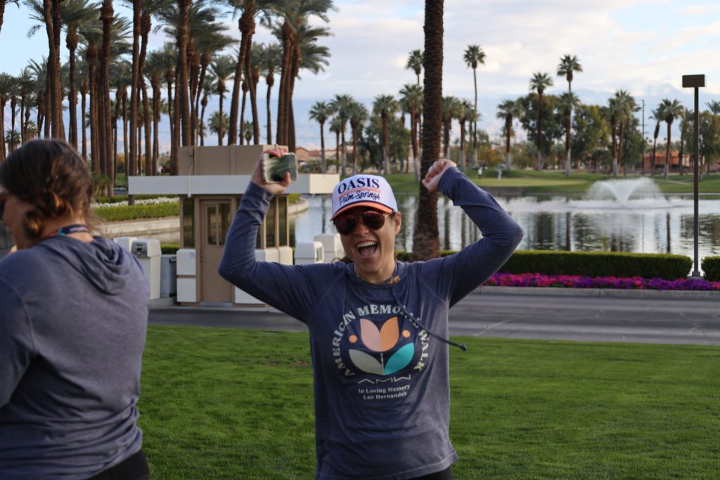 A person in a sweatshirt and cap is joyfully raising their arms, holding a phone. They are outdoors near a palm-lined pathway and a lake with a fountain at the National Sales Conference hosted by ASB Hosts. The sky is partially cloudy, and there are more people in the background.