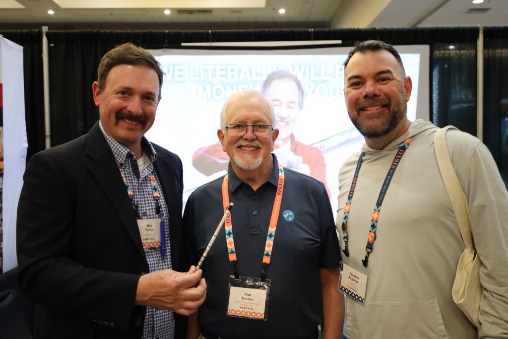 Three men smiling and standing together at the National Sales Conference in Palm Springs, each wearing name tags. One holds a pen, with a booth display in the background showing a message about literacy and money management, proudly presented by ASB Hosts.