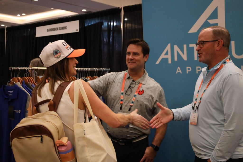 At the National Sales Conference in Palm Springs, a woman with a backpack exchanges a friendly handshake with a man at an apparel booth. Another man stands nearby, smiling warmly. Clothes are neatly displayed on racks behind them, beneath a partly visible blue "APPAREL" banner.