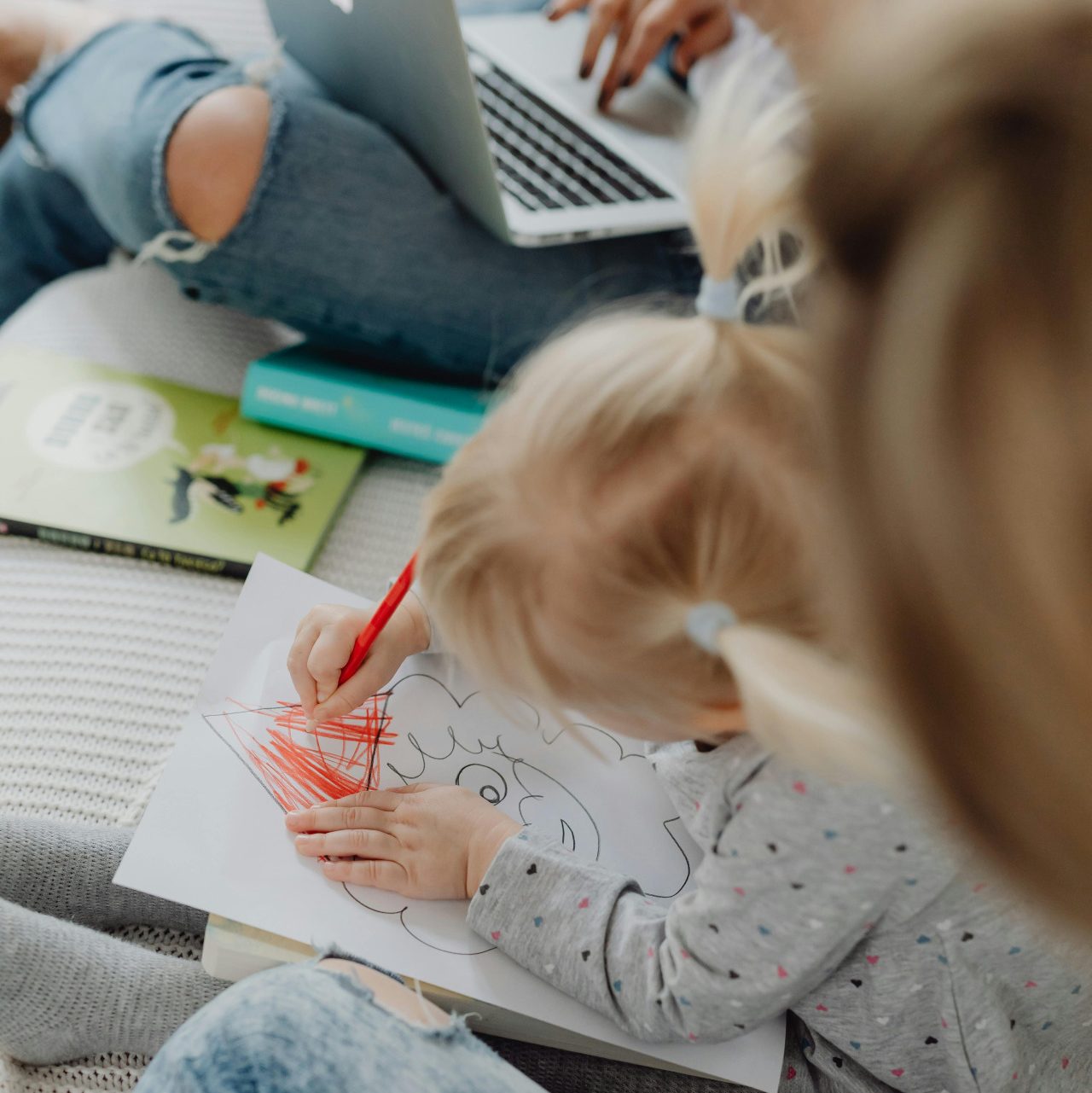 A child enthusiastically colors a drawing with a red crayon while sitting on the floor. Nearby, an adult in a gray sweater and jeans watches patiently. A laptop and books are scattered in the background, resembling a cozy home filled with branded print materials.