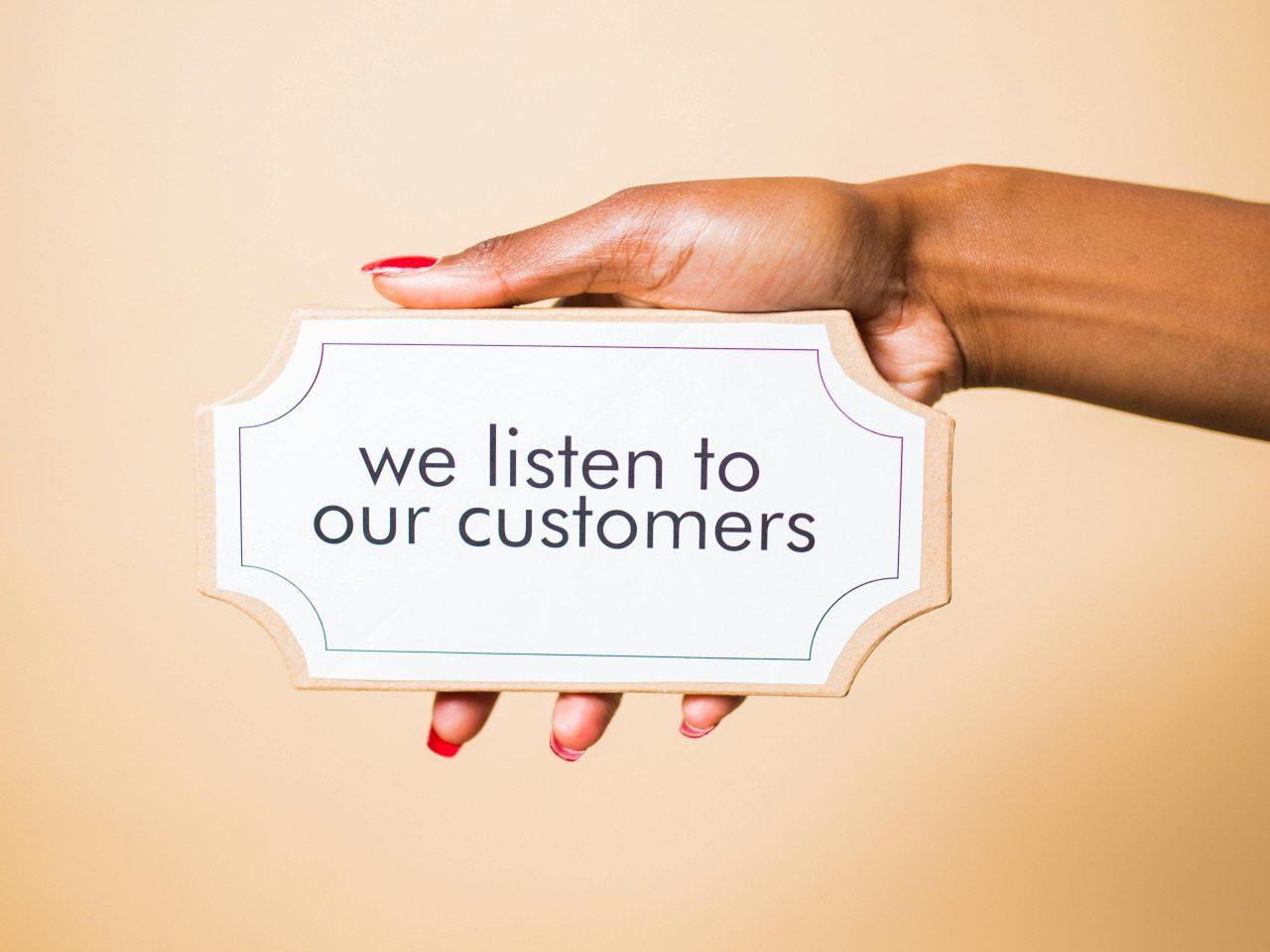 A hand with red nail polish holds a small sign against a beige background, reading "we listen to our customers," highlighting our commitment to patient satisfaction.
