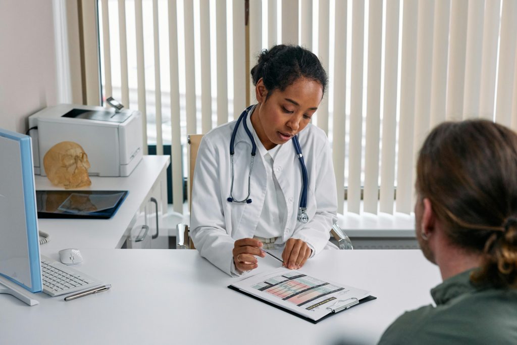 A doctor in a white coat and stethoscope consults with a patient across a desk, aiming to enhance patient satisfaction. The doctor points to a document, as a computer and anatomical model nearby emphasize the professional setting of the medical office with vertical blinds.