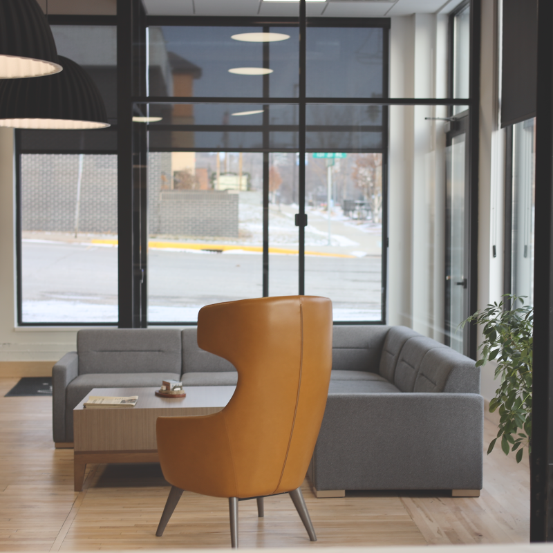 A modern lounge area with a gray sectional sofa, a wooden coffee table, and a distinctively shaped tan leather chair. Large windows let in natural light, revealing a street scene outside. A potted plant sits by the window.