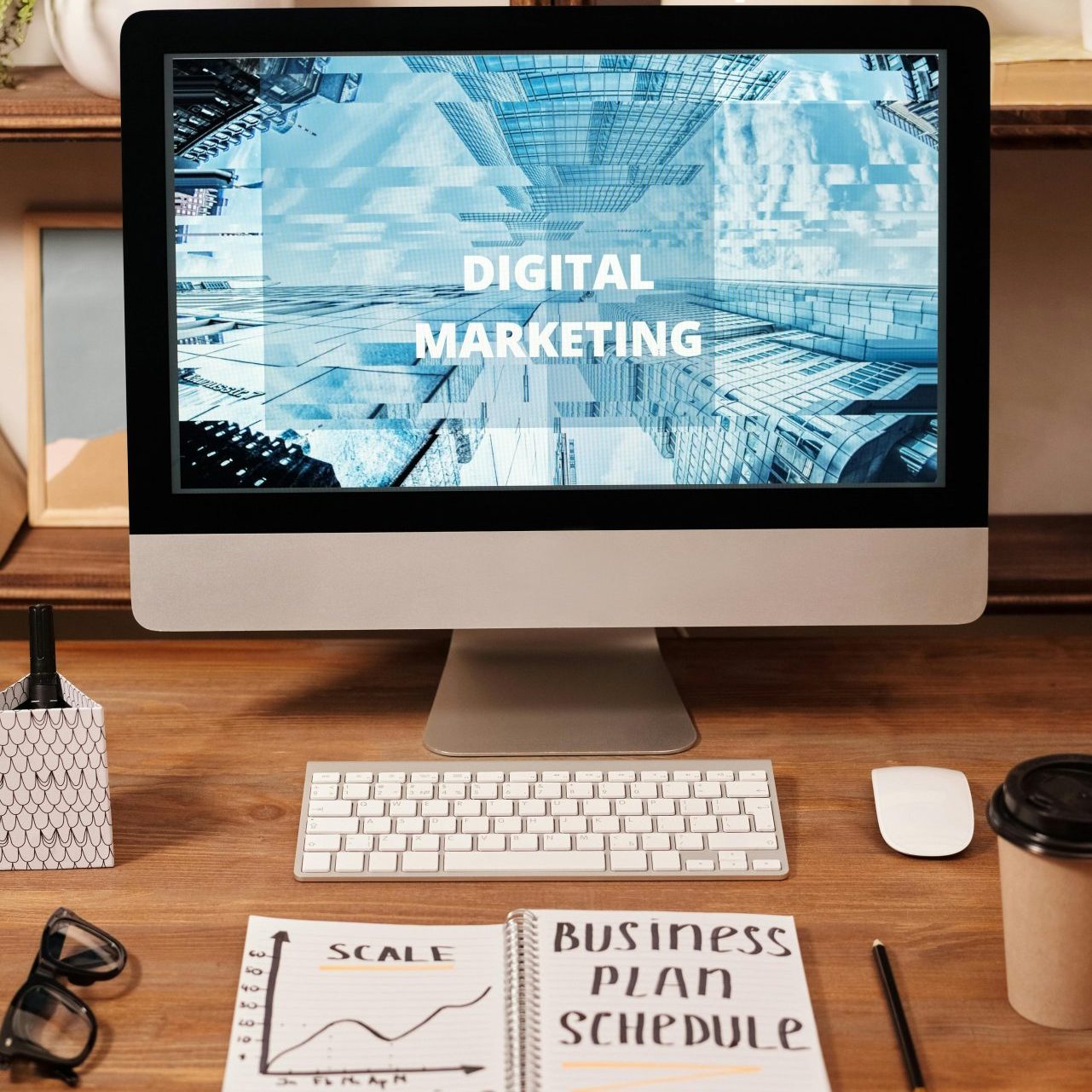 A computer monitor displays "Digital Marketing" on a desk with plants, a notebook labeled "Business Plan Schedule," a pen, glasses, a coffee cup, and a decorative box—perfect for targeting In-Market Buyers with effective Combination Marketing strategies.