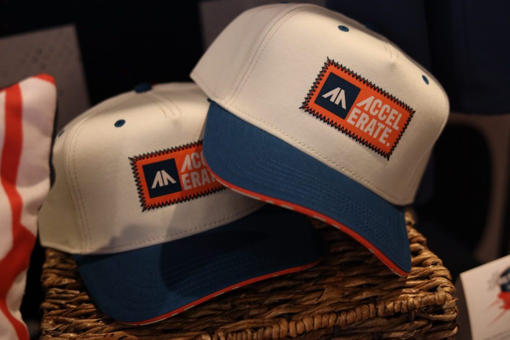 Two white and blue baseball caps featuring the bold “ACCELERATE” logo in orange and blue rest on a woven basket—perfect for showing your ASB pride at the next Accelerate Event in Bloomington MN.