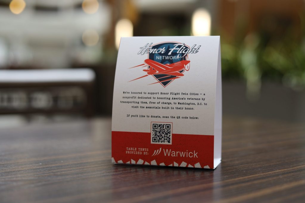 A folded table card displays the "Honor Flight Twin Cities" logo, a brief description about supporting veterans, a QR code, and the Warwick logo on a wooden table with a blurred background at the ASB Accelerate Event.