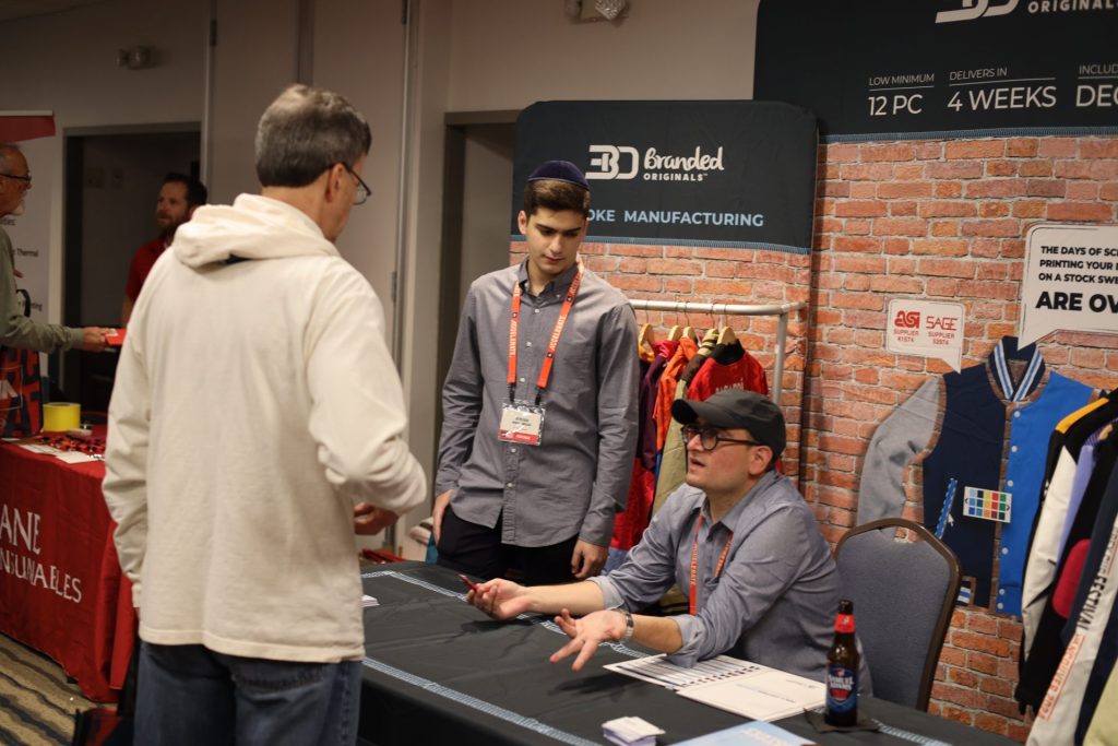 Two men in gray shirts stand behind a table at an Accelerate Event booth, talking to a visitor. Behind them is a display about custom jacket manufacturing. The scene appears to be at a trade show in Bloomington, MN, possibly hosted by ASB.